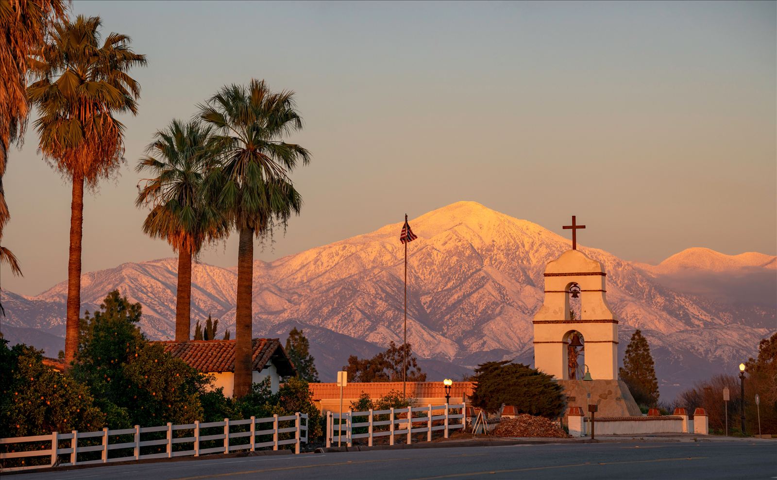 A mission-style bell tower and palm trees framed by a snow-capped mountain at sunset, an iconic Inland Empire view.