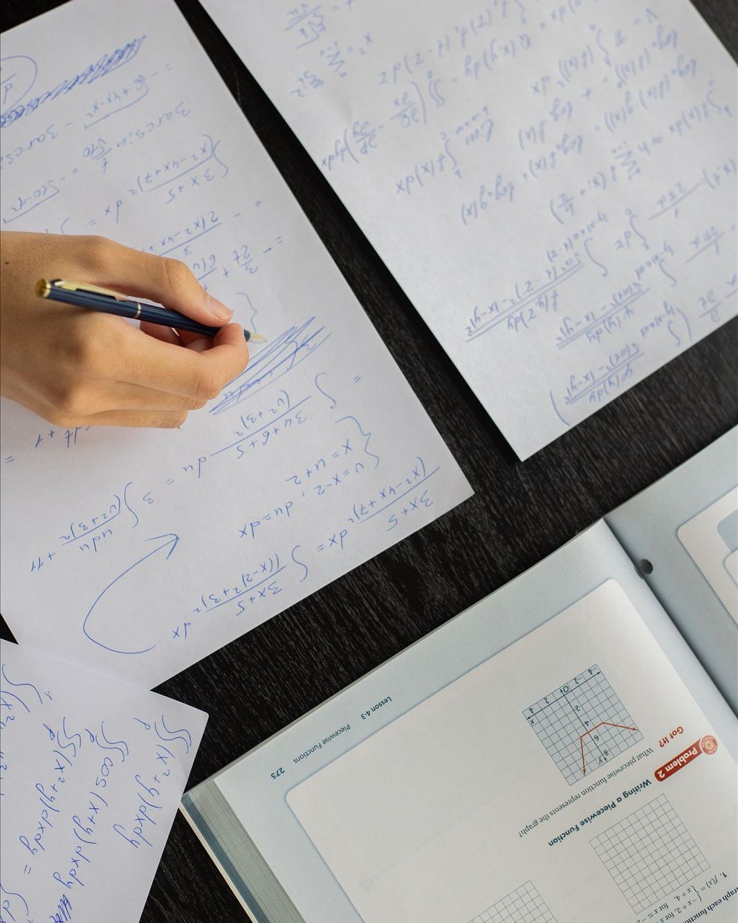 An overhead view of papers covered in handwritten calculus notes, next to an open math textbook.